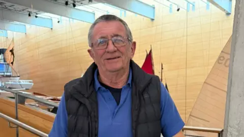 A man with short dark-grey hair, a blue polo shirt and a dark blue body warmer in a room with wooden walls