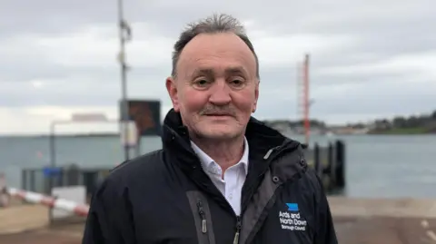 A brown haired man outdoors near a waterfront, wearing a black jacket with the “Ards and North Down Borough Council” logo on it. Behind them are harbour structures, railings, and calm water, with a cloudy sky overhead.
