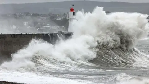 Greg Martin A huge foamy wave crashes into the pier at Newyln Harbour in Cornwall. The lighthouse can be seen beneath the wave, with a beach in the foreground and houses in the background