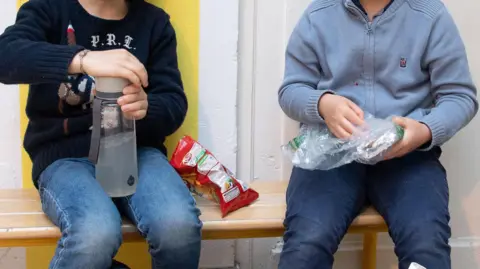 Two young boys are sitting on a bench in a room with one opening a flask of water and he has an open bag of crisps next to him while the other boy is opening a plastic bag which has foil-covered sandwiches inside.