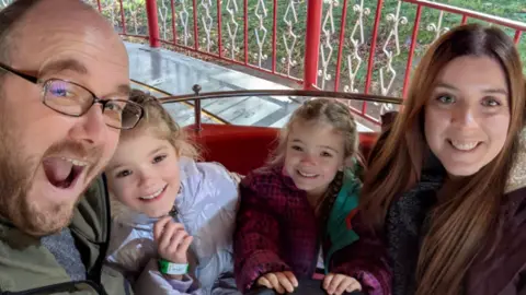 Other A man in glasses, woman with long dark hair and two young girls are all looking at the camera and smiling. The young girls both have blonde hair to their shoulders.