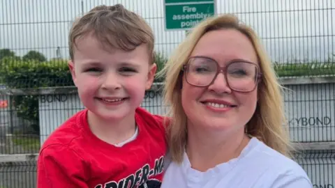 A child and a women standing looking into the camera. The boy on the left, has dark brown hair and is wearing a red t-shirt. The women on the right is wearing a white t-shirt and wearing glasses. 