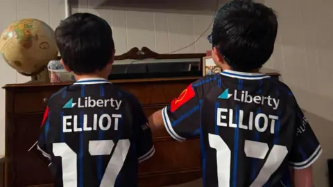 Elliot family Two little boys stand side by side with football strips with Elliot 17 on the back of them