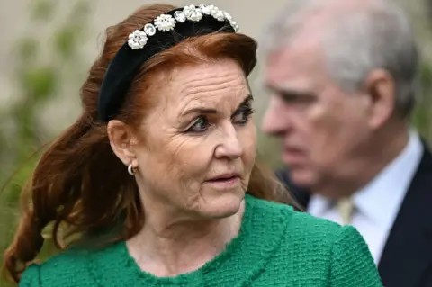 AFP Sarah Ferguson, followed by Andrew Mountbatten-Windsor (formerly known as Prince Andrew, Duke of York), leaves St. George's Chapel, in Windsor Castle, after attending the Easter Mattins Service, on March 31, 2024. Ferguson is dressed in a green collar-less jacket. She has long red hair and is wearing a black headband embellished with pearls. Mountbatten-Windsor is behind her and out of focus.