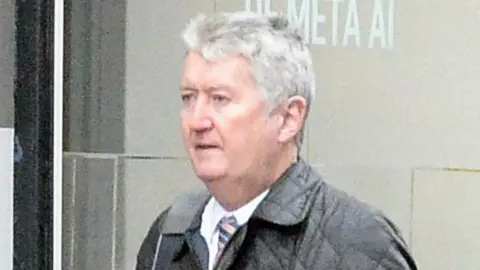 Andrew Hyams, a man with short grey hair wearing a white shirt, striped tie and black padded jacket, walks past a building.