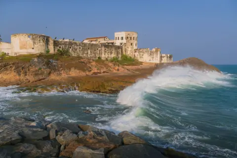 Getty Images A side view of the exterior of Cape Coast Castle with the waves of the Atlantic crashing against the beach beneath the castle's walls.