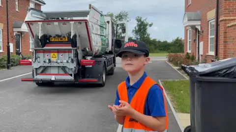 Image shows Ashton stood with orange high vis on and blue top. He has a black cap on that says 'Biffa' in red. Behind him is the food waste truck.