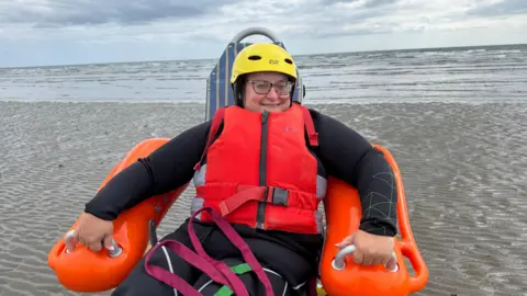 Naomi Herron in a floating chair with orange arms and blue back. She is wearing a black wetsuit, a red lifejacket, and a yellow hat. She is wearing glasses. She is on the beach.
