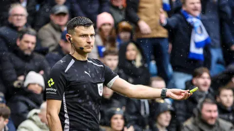 A linesman's outstretched hand holds a yellow tube which was thrown from the Pompey section of the crowd during the Portsmouth-Southampton match on 25.1.26.