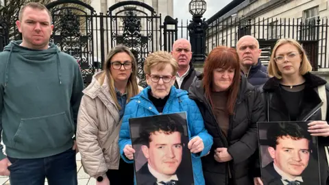 Fergal McCusker's family outside the High Court in Belfast which can be seen in the background behind black iron railings. It is a classical-style building in Portland Stone. There are three men and five women including Fergal's mother Christina who is in the centre in a blue coat, with short blonde hair and red-rimmed glasses. She and the woman on the far right are holding up photos of Fergal who has brown hair and is wearing a formal outfit with a winged collar and cravat.