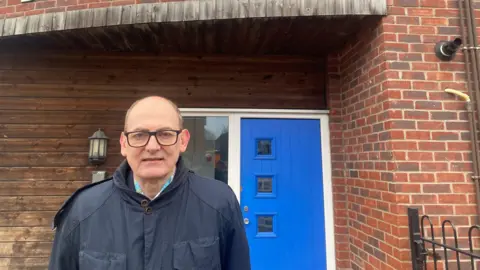 Chief Executive of Elim, Paul Smith, stands outside the front door of Bouverie Court in a navy blue overcoat. He is wearing glasses and is straight-faced.