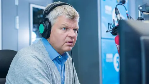 Adrian Chiles is a man in his 50s sitting in front of a microphone wearing a blue shirt and grey cardigan. He has grey short hair and is wearing black and green headphones. He is concentrating and looking at a screen as he talks 