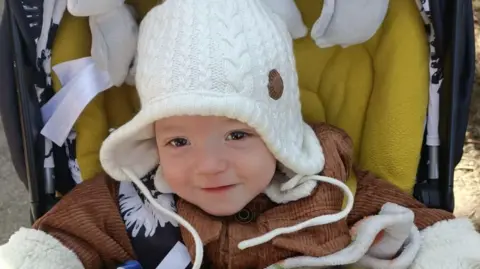 A young boy sits in a push chair and smiles at the camera. He is wearing a white woolly hat and brown jacket