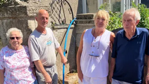 Four residents stand outside a house. There are two women and two men. They are all wearing shorts and t-shirts. One of the men is holding a blue pipe in his hand. Behind them is the stone wall of a house.