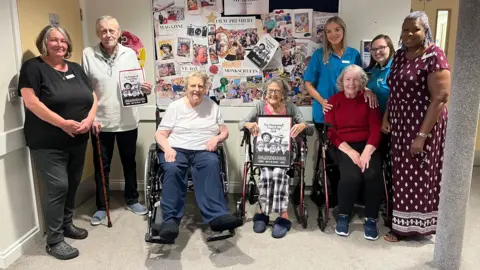 Monkscroft Care Centre A group of elderly care home residents smile as they hold up posters for a movie they have starred in, The Monkscroft Thursday Club, joined by a few carers.