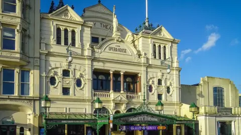 Escenas de la Isla de Man. Un teatro ornamentado de la época victoriana con una fachada color crema. En el tercer piso se lee "Gaiety Theatre and Opera House" (Teatro y ópera Gaiety), junto con una estatua griega de una mujer que alza una antorcha. En la planta baja, sobre la entrada, hay un refugio de acero y vidrio en voladizo.
