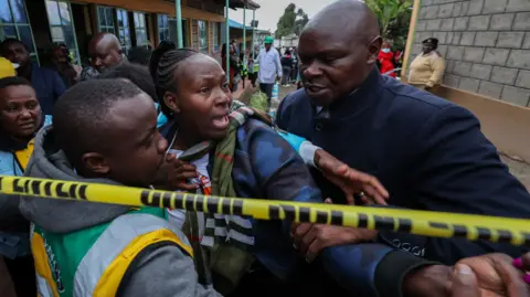 EPA A woman appears visibly distressed as men hold her back in front of police tape at a school in Kenya 