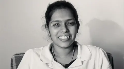 Daisy Moseley Black and white portrait of Cynthia Fernandez. She has dark hair which is tied up and is wearing a beaded necklace and shirt with a collar. She is sat in a chair against a solid background, looking directly at the camera and smiling. 