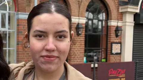 Martin Heath/BBC Ana Aspas looking at the camera with dark hair tied back and a silver earring visible in the right ear. She is wearing a beige coat. There is a brick-built restaurant behind her with a "PanZini" board forward of the entrance.