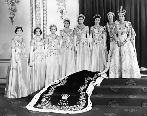 PA Archive Seven women stand in a row to the left of Queen Elizabeth, all wearing white formal dresses and tiaras, with the Queen's velvet robe flowing down a set of stairs infront of them, in a black and white print.