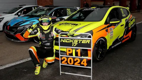 Nicky Taylor Nicky Taylor kneels to the left of a sign saying "P1 2024" to celebrate the Clio Cup GB Championship win. He's in front of his title-winning car, beside two other competing cars.