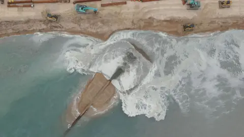Looking straight down on to sea and beach where a pipe is pumping material on to a beach from the sea causing white froth on the edge of the sand.