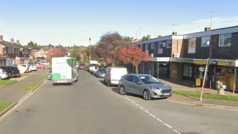 A residential street with cars parked on both sides of the road and a Morrisons van driving between them. Two convenience stores are situated next to each other. It is a sunny day with blue sky. 