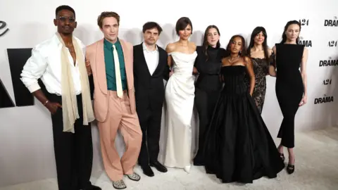 Getty Images Mamoudou Athie, Robert Pattinson, Kristoffer Borgli, Zendaya, Alana Haim, Jordyn Curet, Zoë Winters, and Sydney Lemmon attend the Los Angeles premiere of A24's The Drama. They are standing on a white carpet in front of a white background with "The Drama" branding on it. They are all dressed in black and white formal wear except Pattinson, who is wearing a pink suit, green shirt and white tie. 