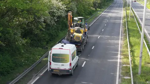 Joe Weir / BBC View of the lorry with a lopsided digger on its back parked on the A63 with a van behind it 