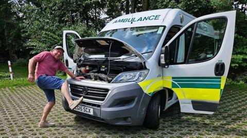 Terry Anderson wearing grey cap has one foot on the radiator of a parked ambulance