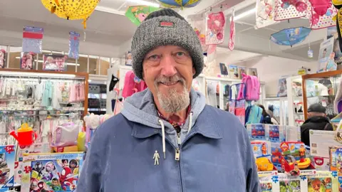 John Molson - a man with a grey beanie, blue jacket and grey hoodie smiling into the camera. He has a grey beard and colourful jigsaw puzzles and children's clothes can be seen in a shop behind him.