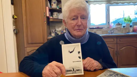 A lady with short brown hair and blue eyes, wearing a blue turtleneck jumper, holds a black and white photograph of a girl in a black bathing suit diving. 