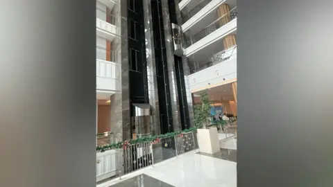 Collette Kerry A hotel lobby with its two lift shafts, made of marble and glass. There are several balconies visible which are white and have glass barriers in front of them.