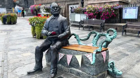 A metal statue of Victor Hugo - an old man with a beard in a three-piece suit, reading a book. He is sitting on a bench, with space for one person to sit next to him, with the back and side made out of a copper-blue octopus and its tentacles. Behind him is a granite town square and an old church, with benches and railings. A man in a bucket hat and T shirt sits on one of the benches. 