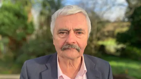 Michael Gallagher. He has white hair with grey moustache, wearing a dark grey blazer and pink shirt. Trees are blurred in the background.
