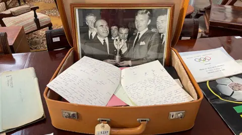 An open light brown suitcase with a photo of Sir Alf Ramsey and Bobby Moore inside, alongside other documents.