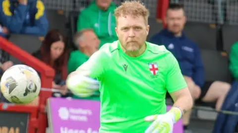A man with brown hair is wearing gloves and a green top. He is in the centre and a ball is nearly halfway up the image on the left. People watching in the background are blurred.