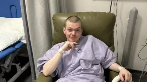 Aidan McAllister A man wearing a purple hospital gown sits next to a bed