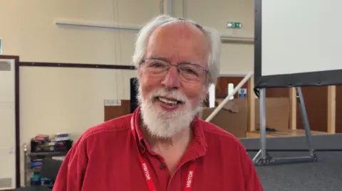 Man with grey hair and beard, wearing red shirt smiling at the camera, stood in front of a stage and presentation board.