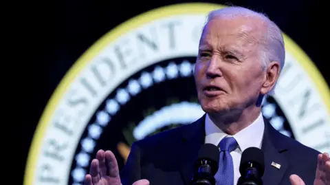 Joe Biden, dressed in a dark suit with a blue tie and white shirt, stands before a microphone, lifting his hands, in front of a seal that reads President of the United States.