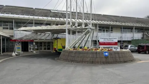 A white sign saying "Emergency Department and Radiology". A smaller white sign underneath which says "MRI". There is a small roundabout outside of the  entrance of the building. There is a yellow and green ambulance outside of the doors. 