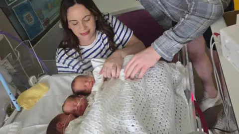 A photo of a woman looking down at three babies who are sleeping. She has got a white top on with blue stripes. 