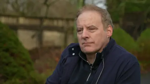 Michael Helps, who is wearing a navy jacket, is staring off camera while in a park in this still from the interview footage. 