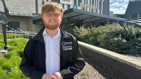 A man with short ginger hair and ginger beard standing in front of Telford bus station. He is wearing a white shirt and black jacket. The sun is shining in the background with some clouds in the sky behind the grey and white bus station. 