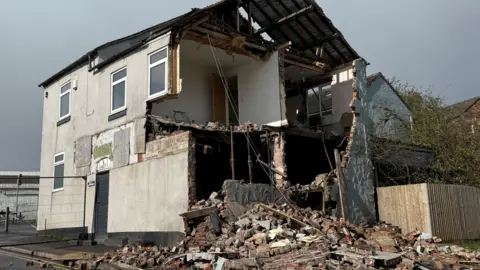 The partially-demolished shell of a building with top floor and roof area exposed. There is a pile of rubble at the base of the building and other buildings visible in the background.