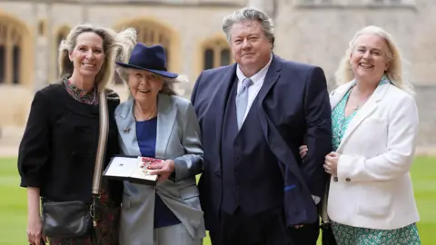 PA Dame Jilly Cooper holds her DBE as she stands with her daughter, son and daughter-in-law on the lawn outside Windsor Castle. Dame Jilly is wearing a navy fedora and a light blue suit with a navy top underneath. She is smiling widely as are all the others in the picture.
