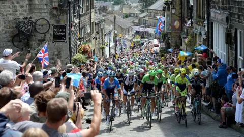 PA Media A huge crowd of cyclists battling uphill in Haworth, Yorkshire. The narrow, cobbled streets are full of riders and spectators who are spilling onto the road. Many are holding up cameras and union flags.