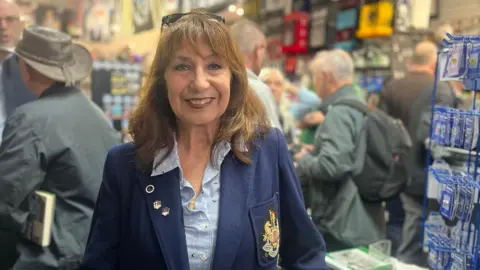 Elizabeth Robledo, the daughter of Chilean footballer George Robledo stands in front of shoppers at the Back Page book shop in Newcastle. She is dressed in a light blue shirt and is wearing her fathers navy blue jacket which has the Newcastle United crest stitched into the pocket. 