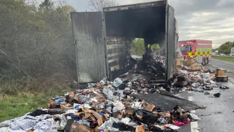 A burnt out lorry with its back doors open on the A1. Items spill out from the lorry onto the road. A fire engine is in the background.