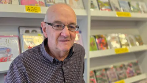 Paul Moseley/BBC Ray Harvey is wearing a patterned shirt, with small red, black and white checks on it and standing in front of a display of greetings cards.